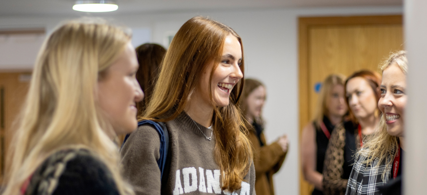 A student standing in a corridor, smiling at a member of staff on the right of the camera.